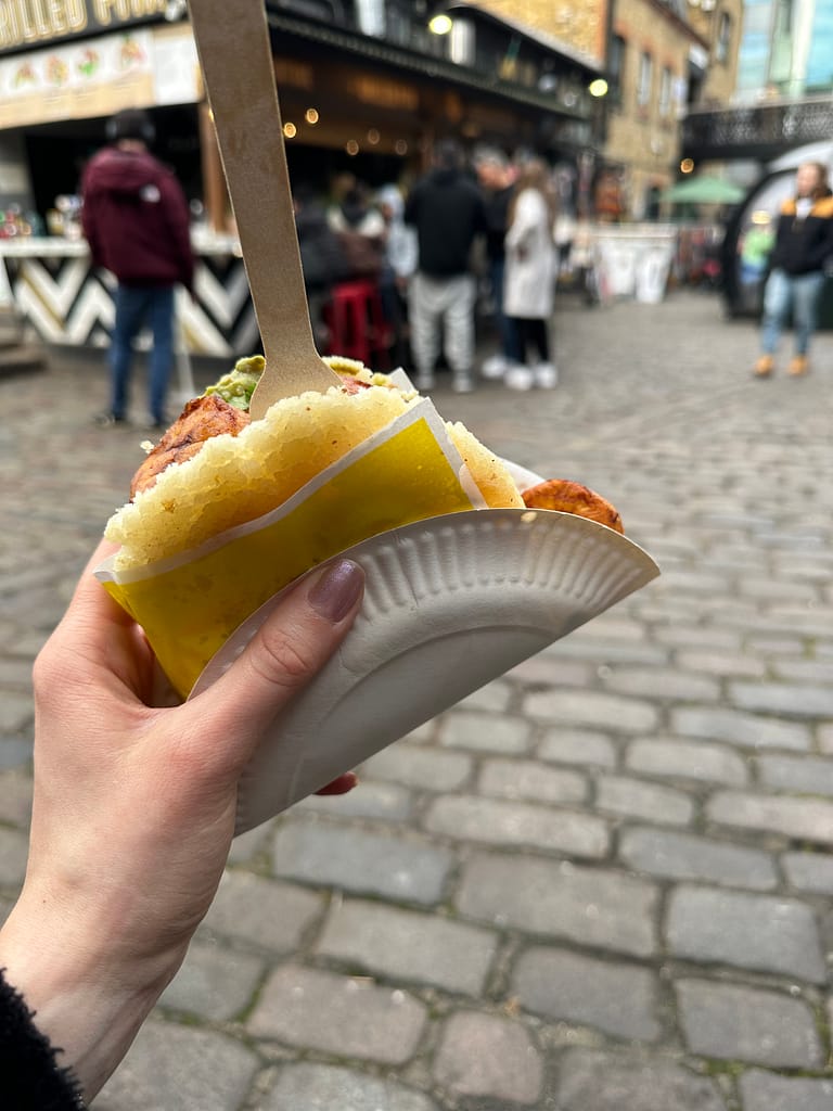 Henkilön kädessä on street food henkinen ruoka, taustalla markkinat